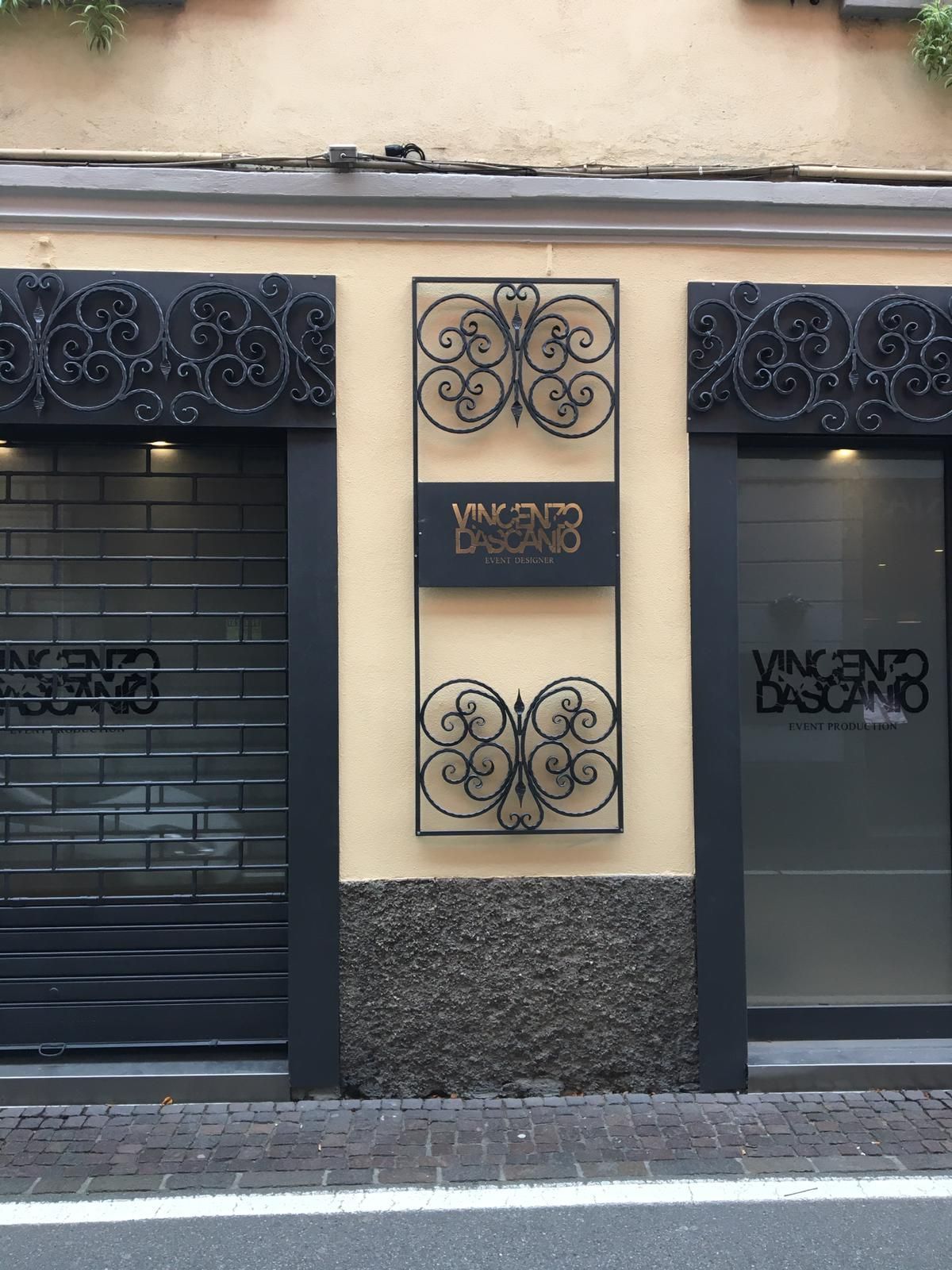 Negozio al piano terra con cancelli in metallo nero, elementi decorativi in ​​ferro battuto e insegna centrale su una facciata beige.