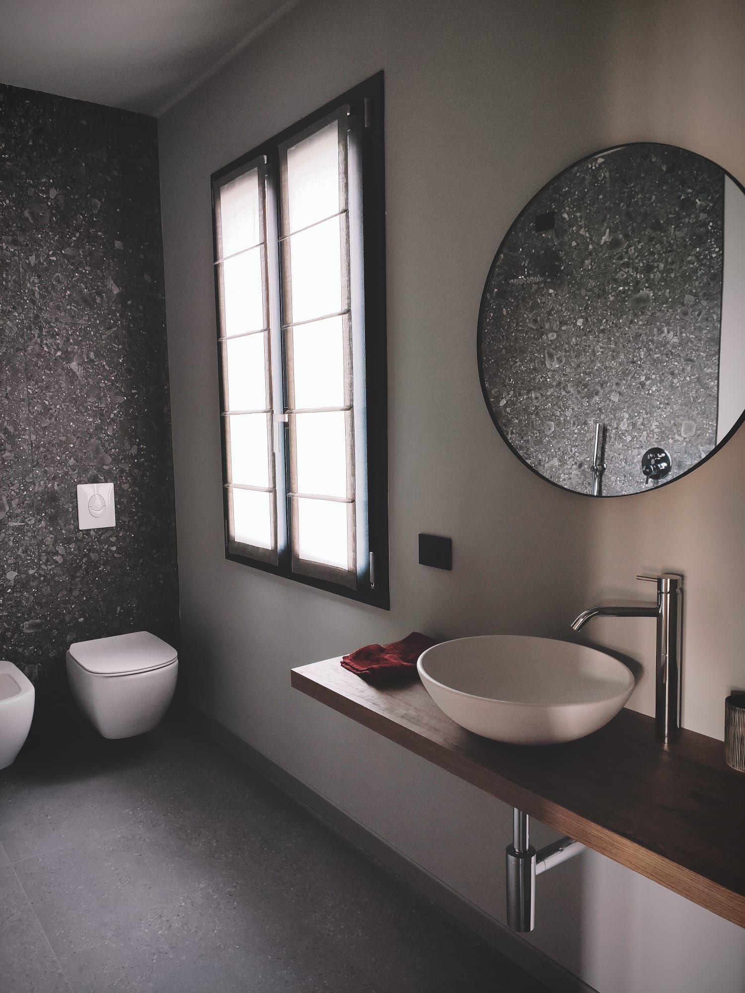 Bagno moderno con pareti scure, finestra, lavabo a ciotola, specchio rotondo e WC sospeso.