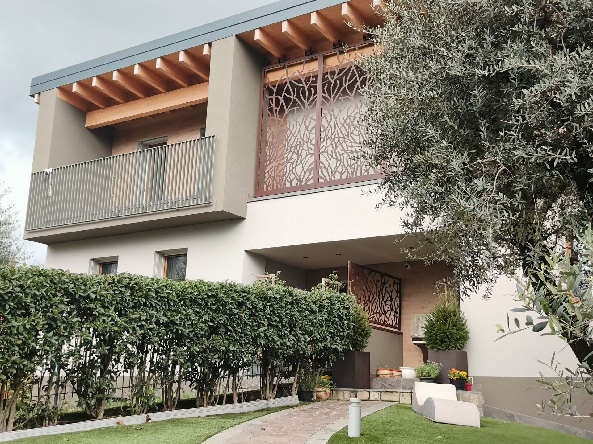 Casa moderna a due piani con balcone, pergolato in legno e siepi rigogliose nel giardino anteriore.