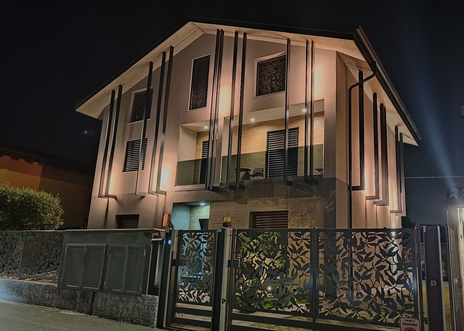 Vista notturna di una moderna casa a due piani con balconi, facciata illuminata e un cancello metallico decorativo sul davanti.