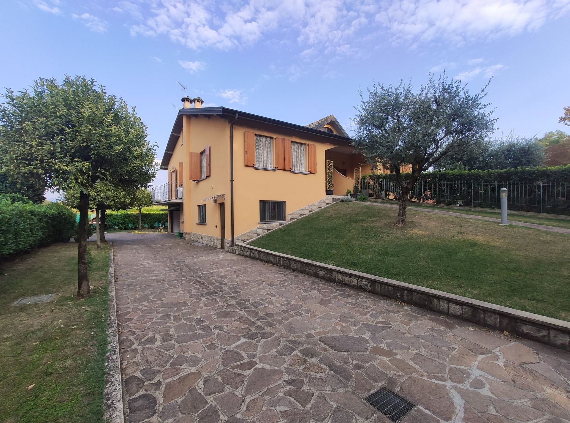Casa di campagna con persiane arancioni accanto a un vialetto in pietra, pendio erboso e alberi sotto un cielo parzialmente nuvoloso.