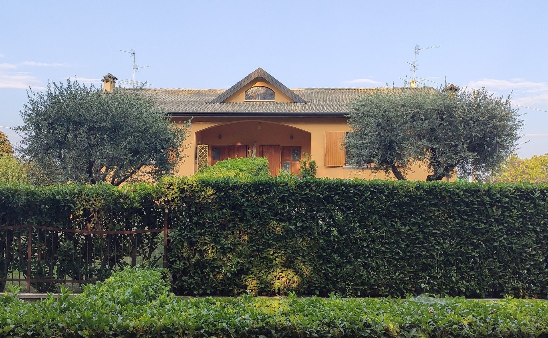 Casa gialla con tetto di tegole rosse dietro un'alta siepe verde e alberi potati.