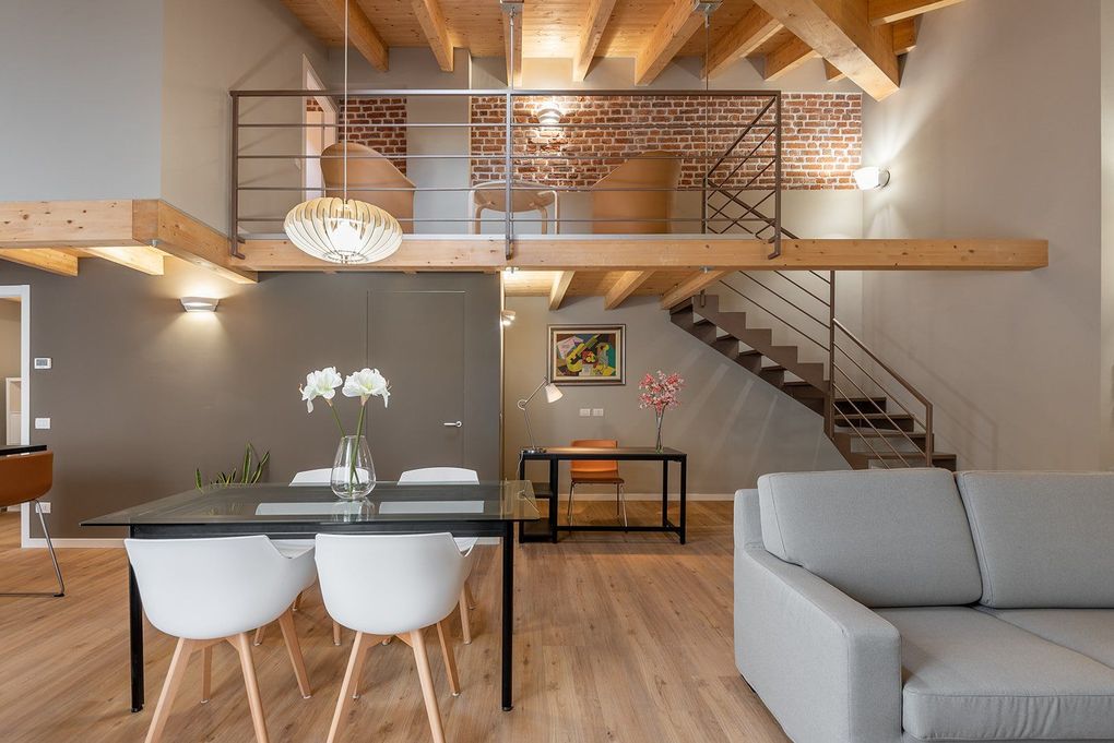 Soggiorno e zona pranzo in stile loft moderno con scala, travi a vista, divano grigio e mobili in legno.