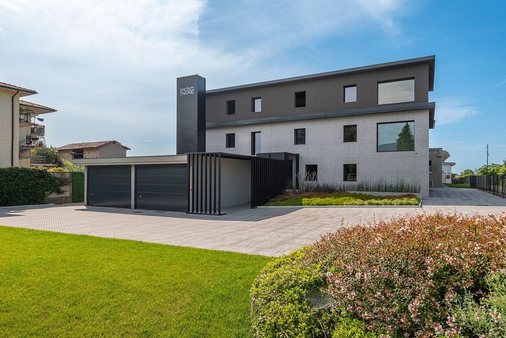 Moderno condominio bianco e grigio con posto auto coperto e prato verde sotto un cielo azzurro.