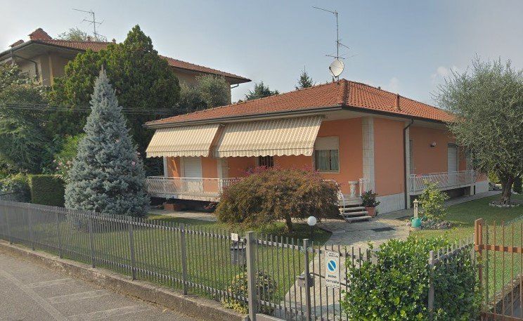 Casa di periferia con tetto rosso, tende da sole a righe e cortile anteriore recintato adiacente a una strada.