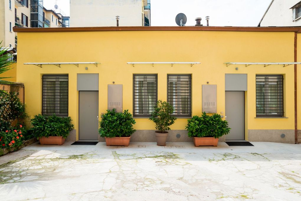 Facciata gialla dell'edificio con tre porte, finestre con sbarre e arbusti in vaso su un patio soleggiato.
