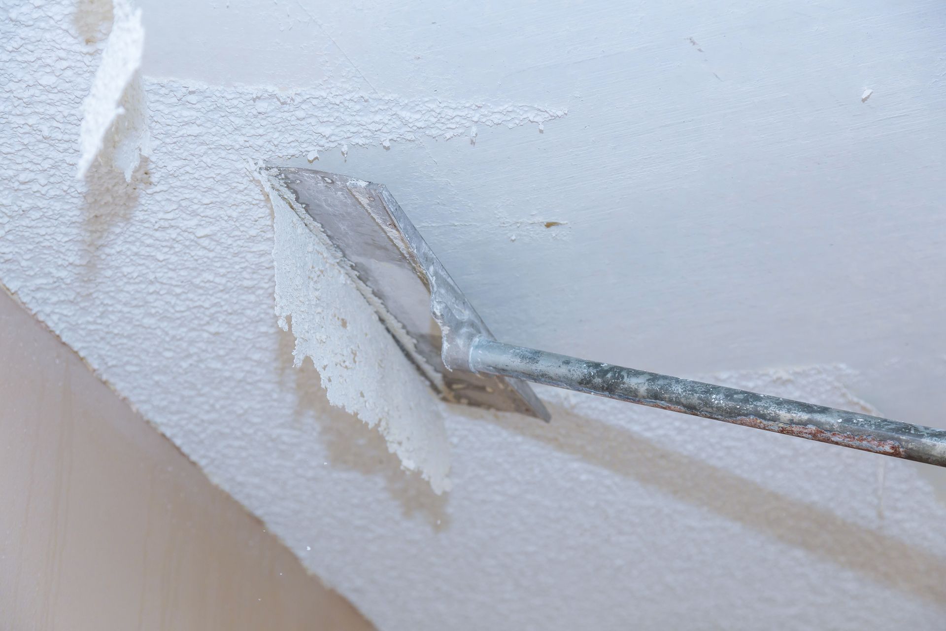 A long-handled scraper removes white popcorn texture from a ceiling, revealing smooth drywall underneath.