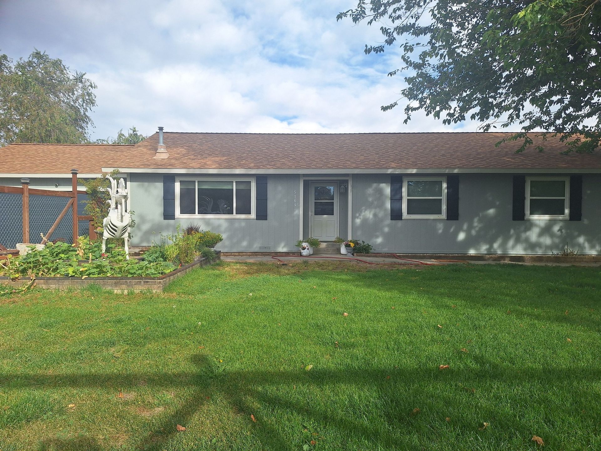 A single-story gray house with a tiled roof, white trim, dark shutters, and a garden bed in the front yard.
