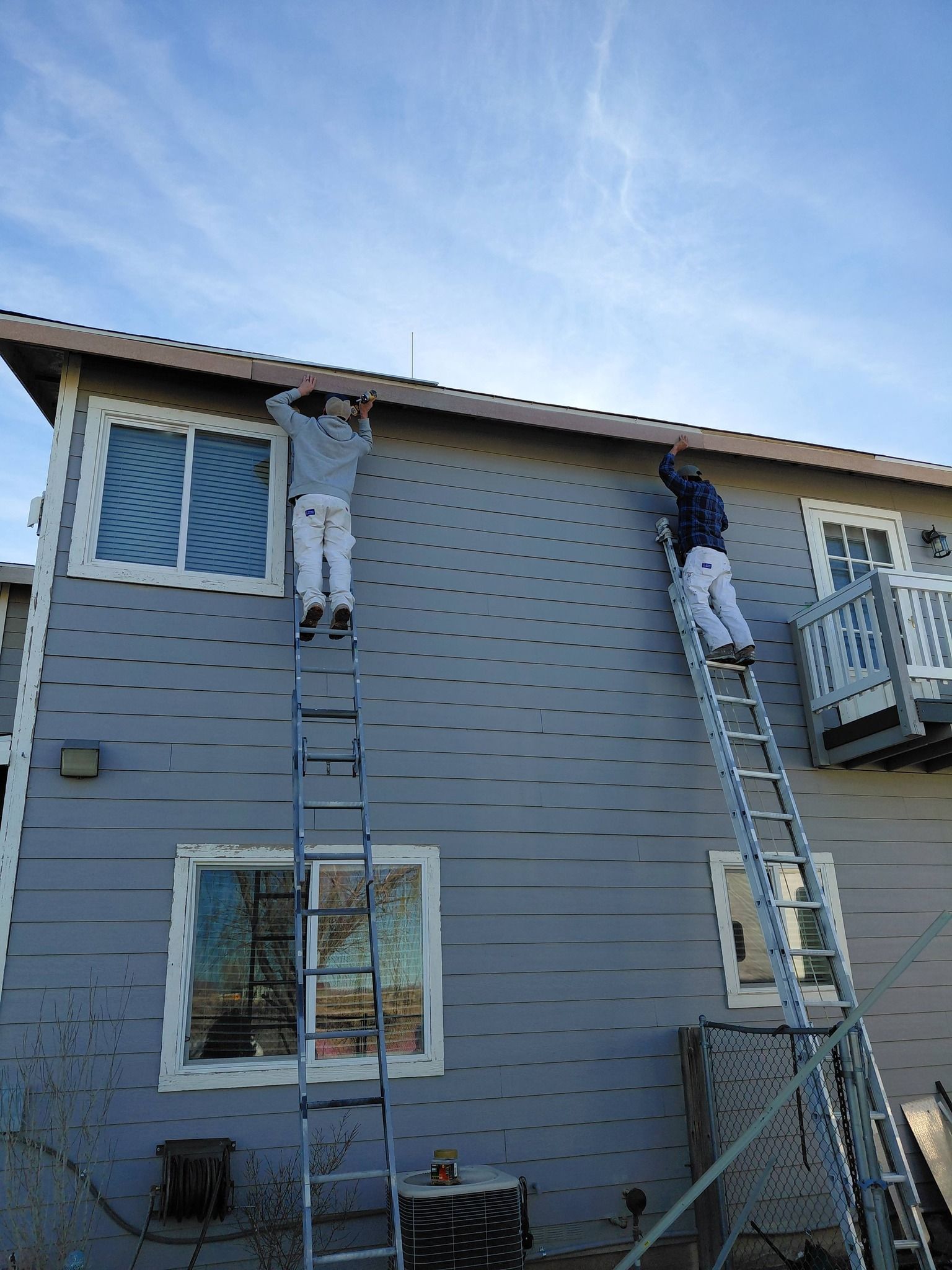 Two workers on ladders paint the grey exterior wall of a two-story residential building.