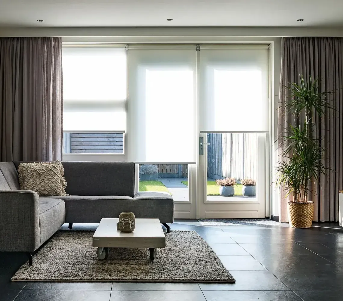 A modern living room with a grey sectional sofa, coffee table, rug, and large windows with curtains and roller shades.