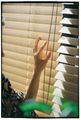 A person's hand reaches out to touch light-filtering wooden blinds with a beaded chain hanging in front of them.