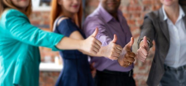 Four people in business casual attire stand together, each extending a hand with a thumbs-up gesture.