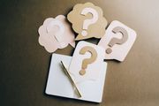 A notebook and pen surrounded by four paper speech bubbles, each featuring a large question mark, on a dark background.