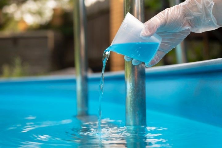 Gloved hand pouring bright blue pool test liquid into a swimming pool