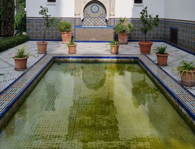 Mosaic-tiled courtyard fountain with green water, potted plants, and ornate tilework backdrop