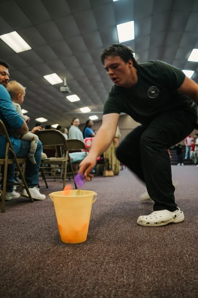 Man in a black shirt crouches to pour liquid into an orange bucket at an indoor event