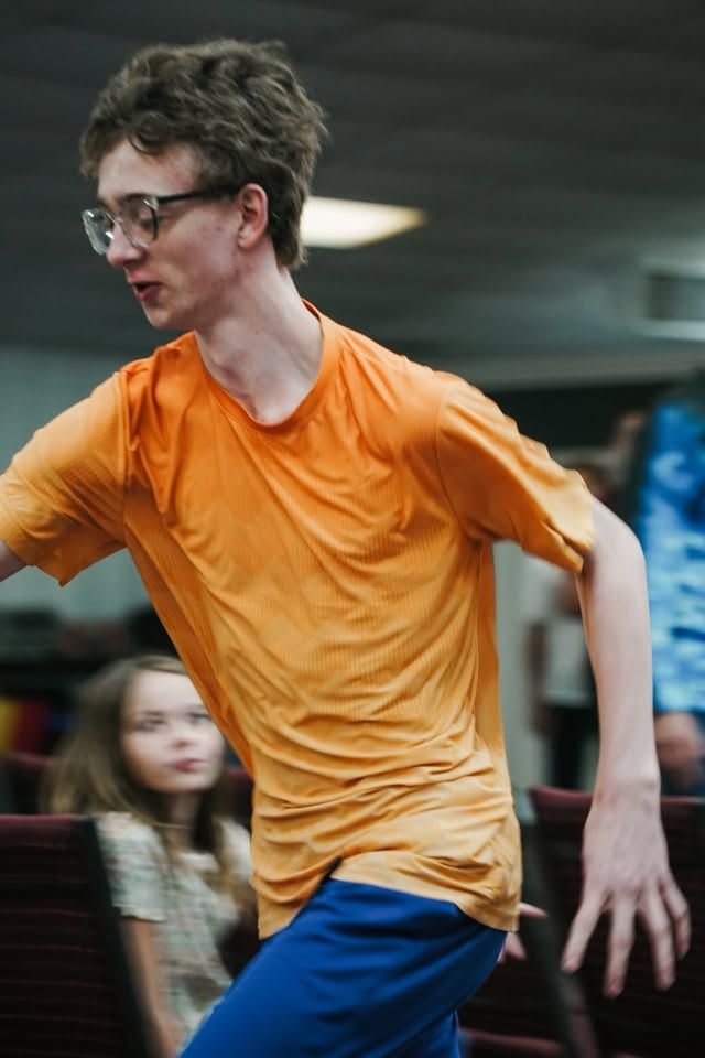 Young person in an orange shirt and blue pants moving quickly indoors, with a blurred crowd behind