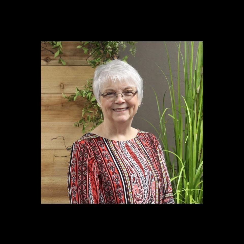 lady with dimples and glasses; gey hair standing in front of a wooden accnt wall, with greenry