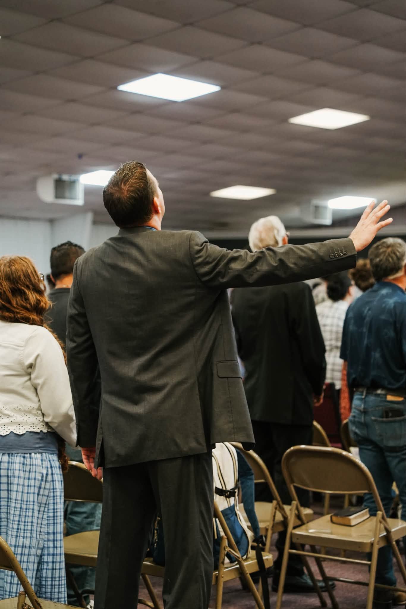 People standing with arms raised in a crowded indoor meeting room with folding chairs