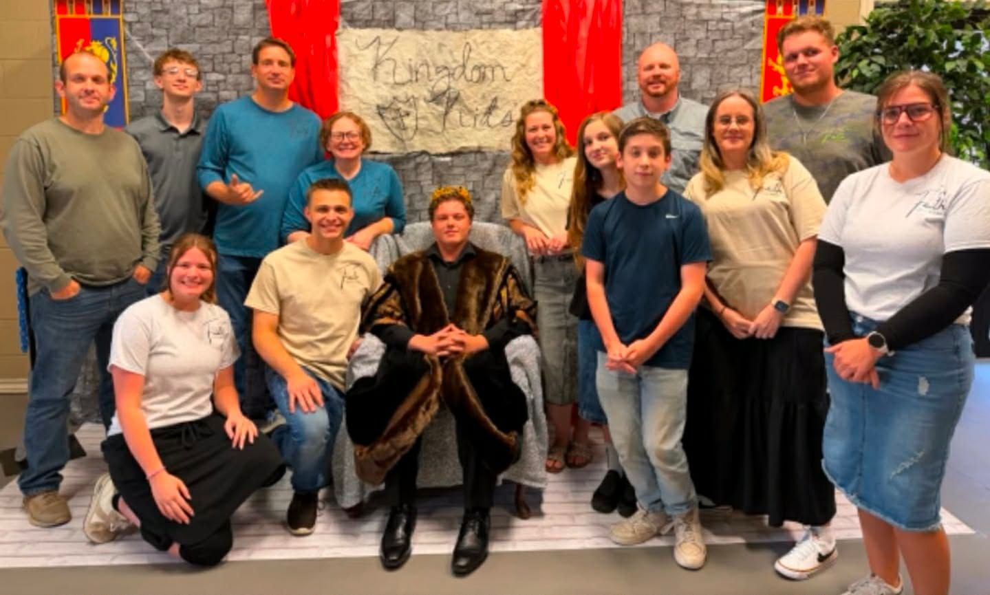 Group of people posing indoors on a stage, with red curtains and a brick wall backdrop.