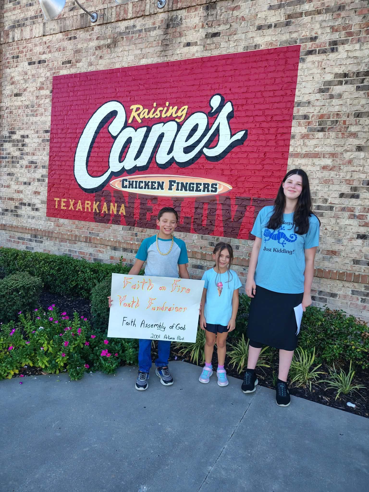 Three people posing by a Raising Cane's sign, with one holding a decorated sign in front of the restaurant.