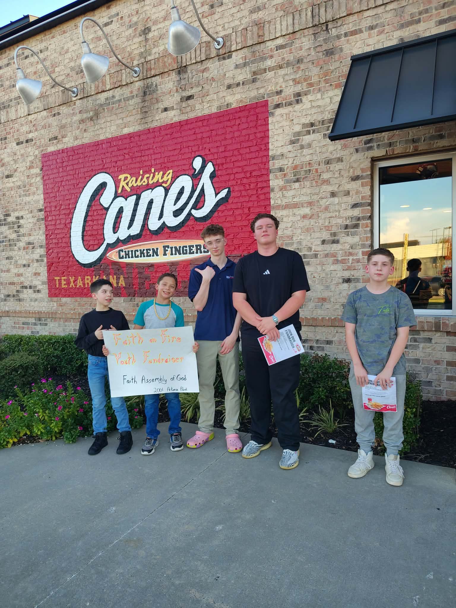 Five people posing outside a Raising Cane’s restaurant, holding signs and folders.