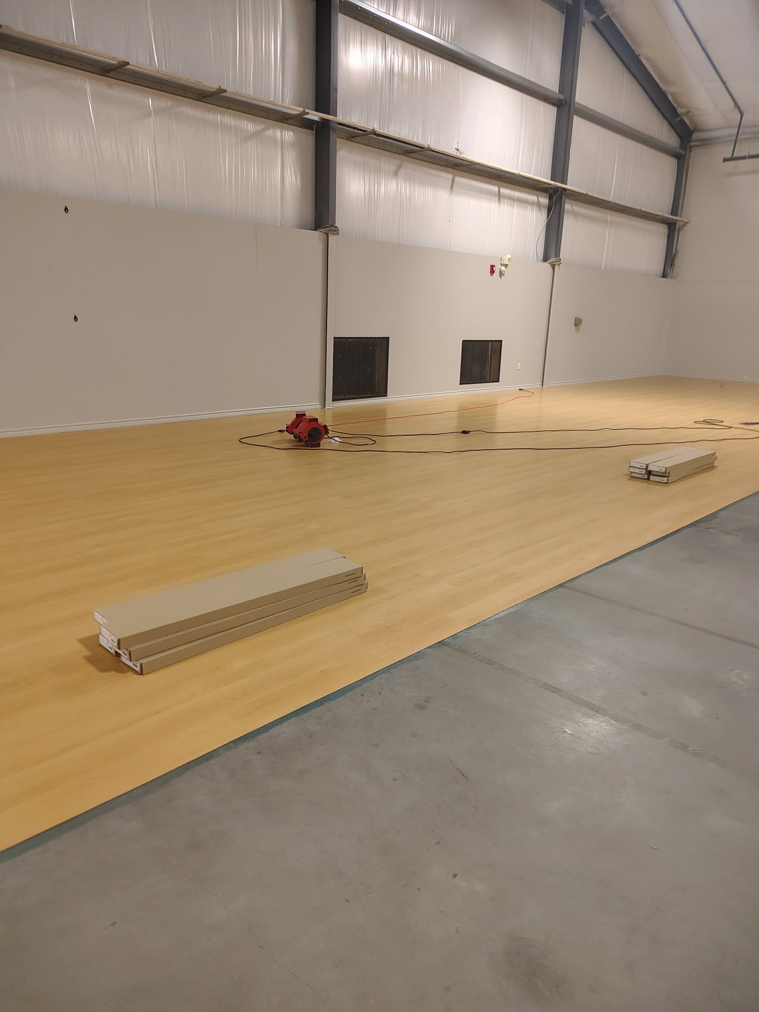 Empty warehouse with a light wood floor, loose planks, and scattered tools under industrial lighting