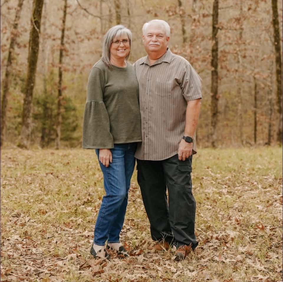 Two people standing together in a wooded autumn setting, smiling at the camera.