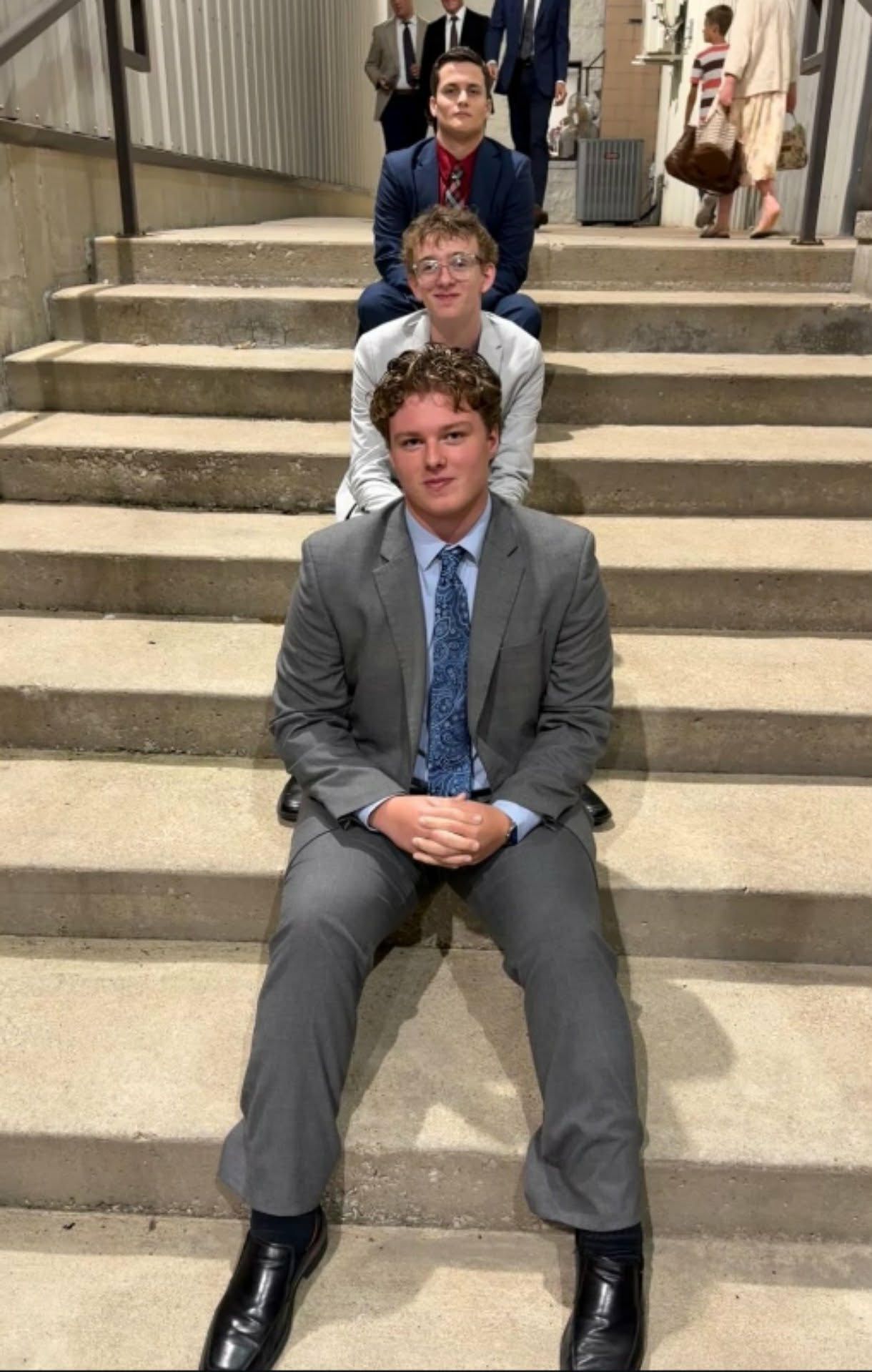 Three people sit on stone steps, with a man in a gray suit in the foreground.