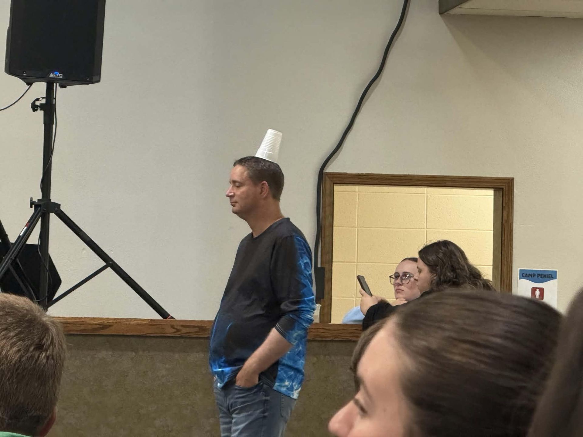 Man standing indoors near a speaker, with seated people in the background and a projection screen nearby