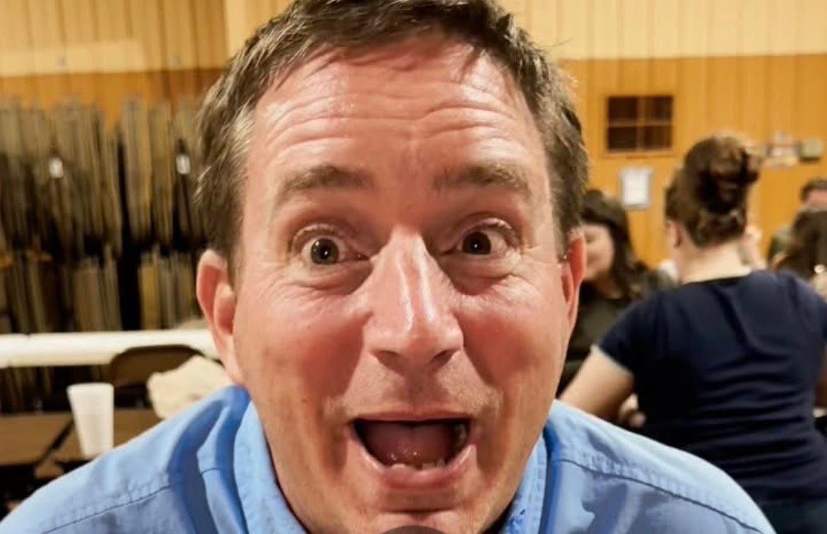 Man grinning at camera in a room with folded chairs and another person in the background
