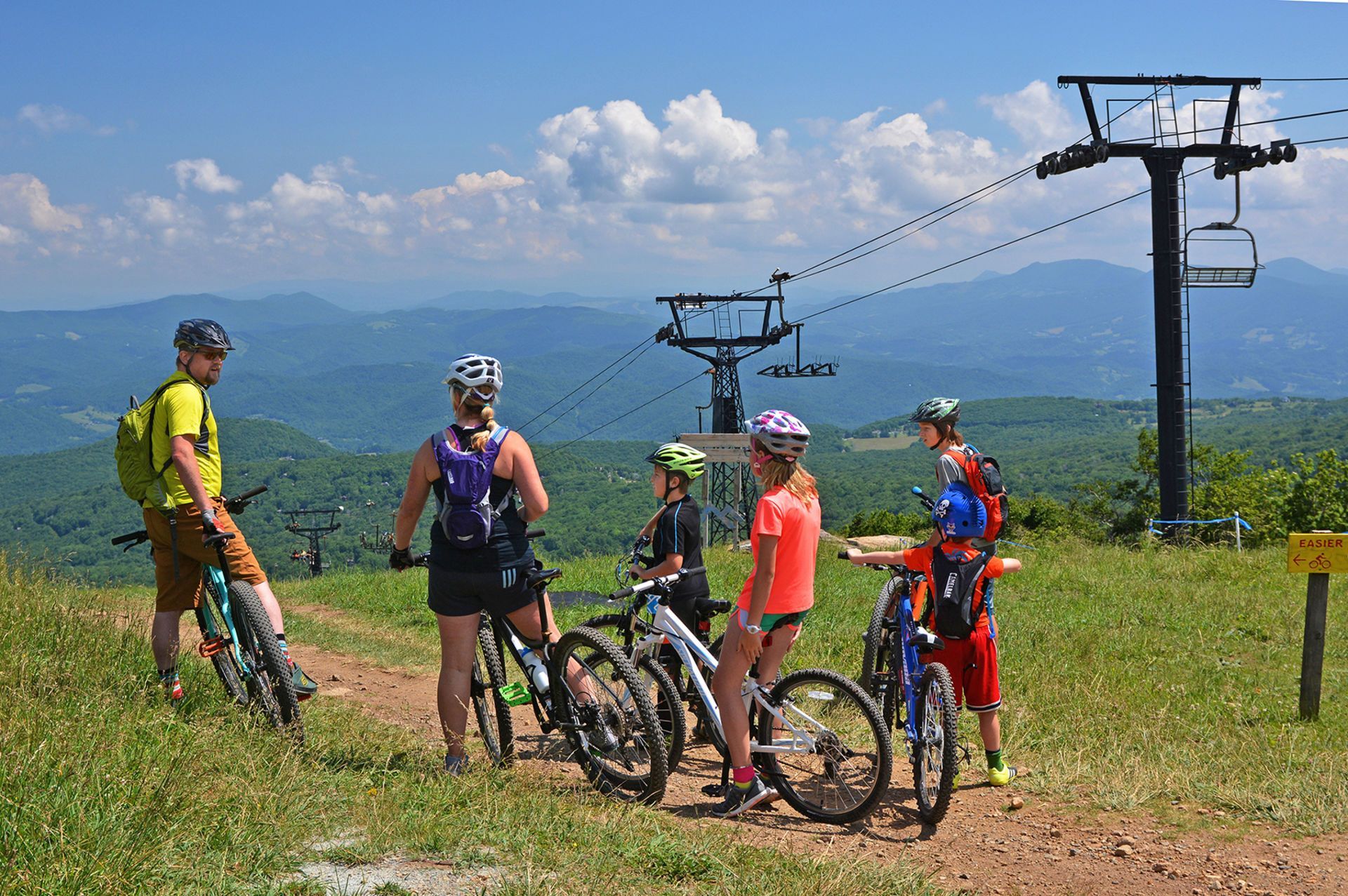 A group of mountain bikers pauses on a grassy hilltop near a ski lift with a scenic mountain vista.
