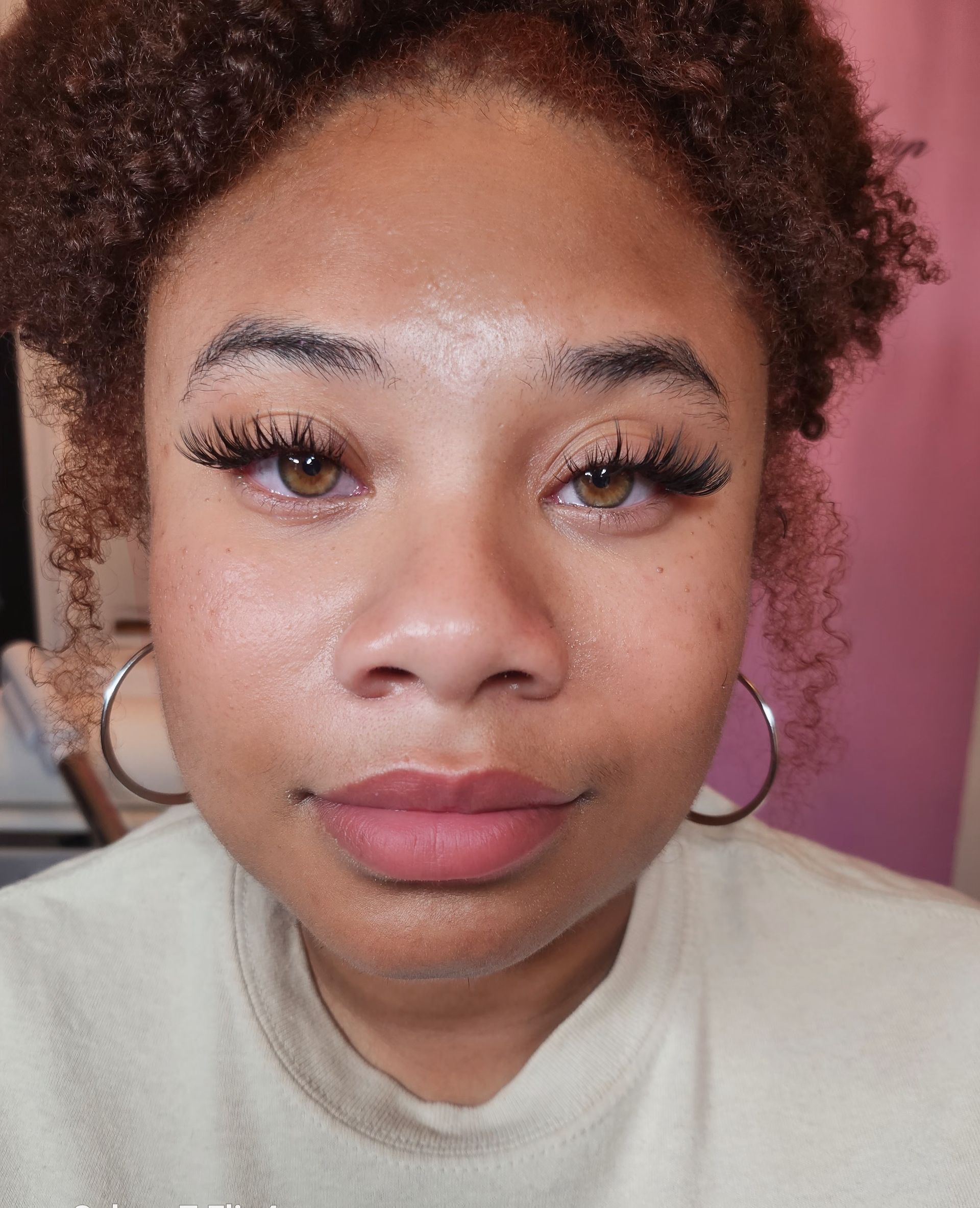 A young woman with curly brown hair and large hoop earrings gazes at the camera, wearing long eyelashes and nude lipstick.