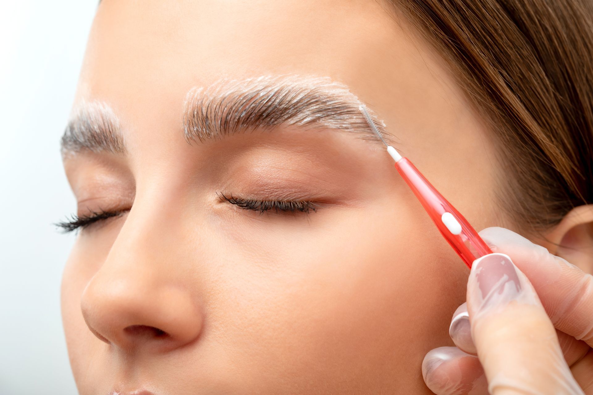 Person having eyebrow makeup applied with a small red brush while eyes are closed