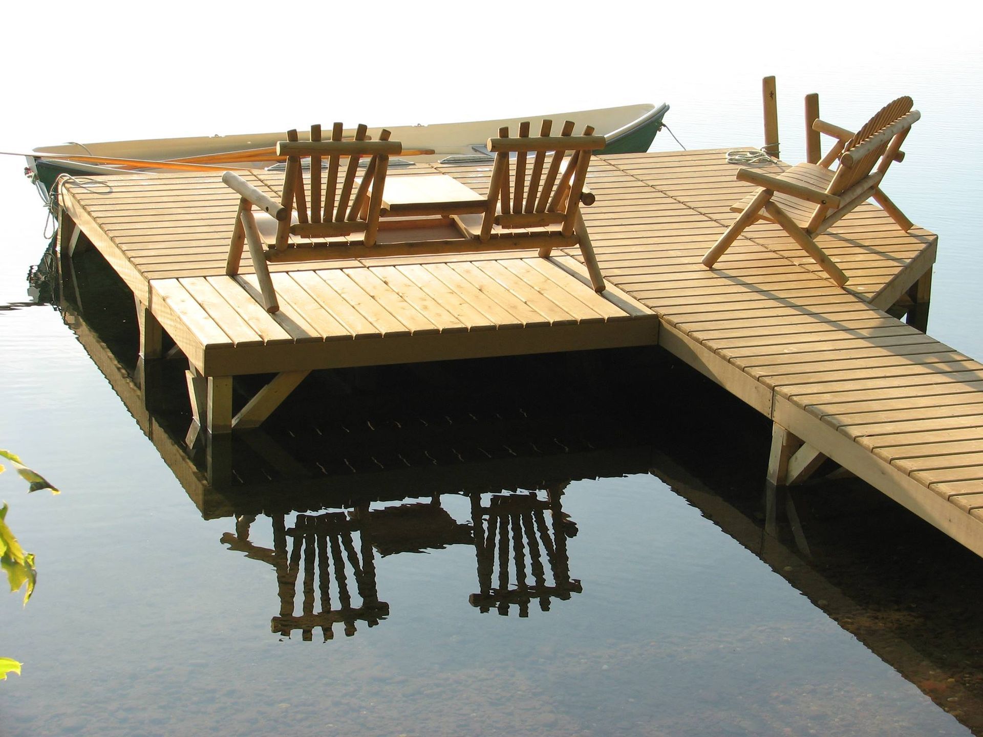 Wooden dock on a calm lake with three chairs and a boat, reflecting in the water.