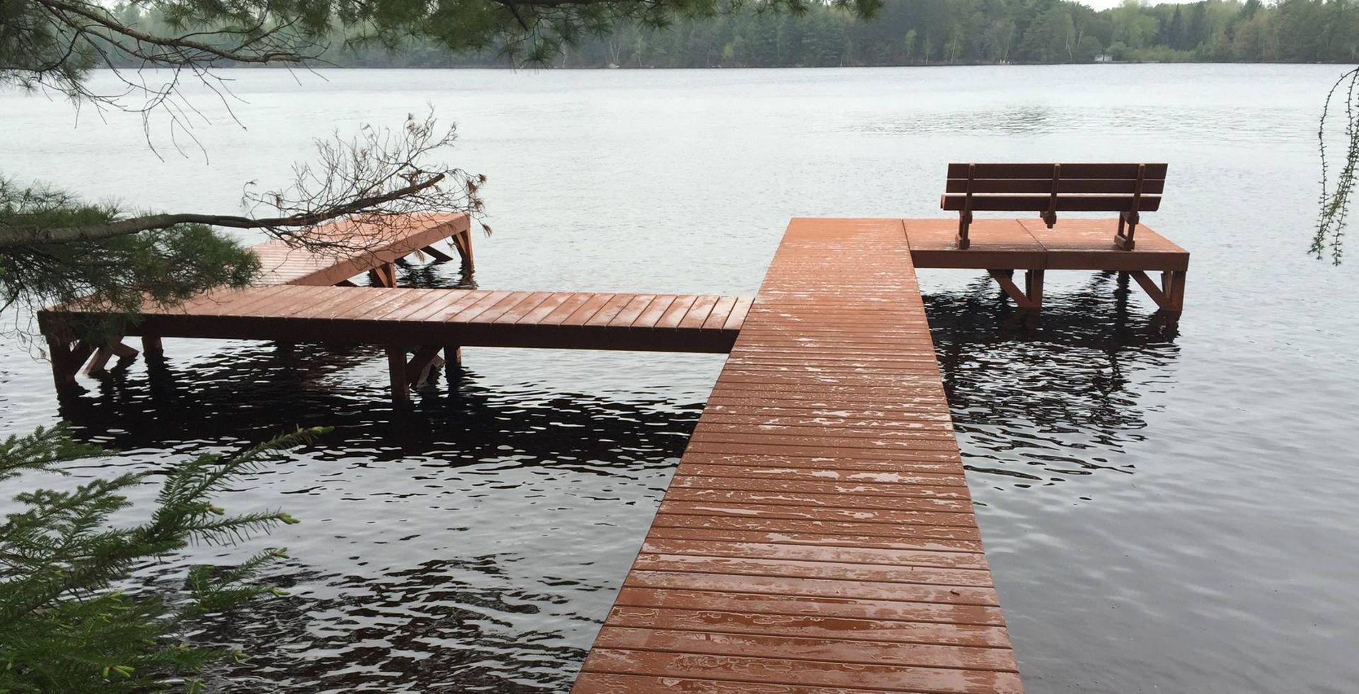 A wooden L-shaped dock extends into a calm, overcast lake, featuring a bench at the far end under overhanging trees.