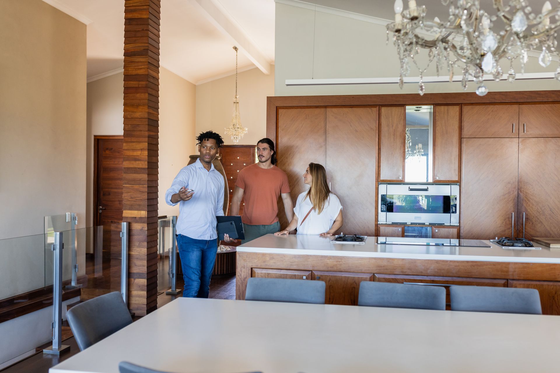 Three people in a modern kitchen. One gestures, others observe near the counter. Wooden cabinets and chandelier.