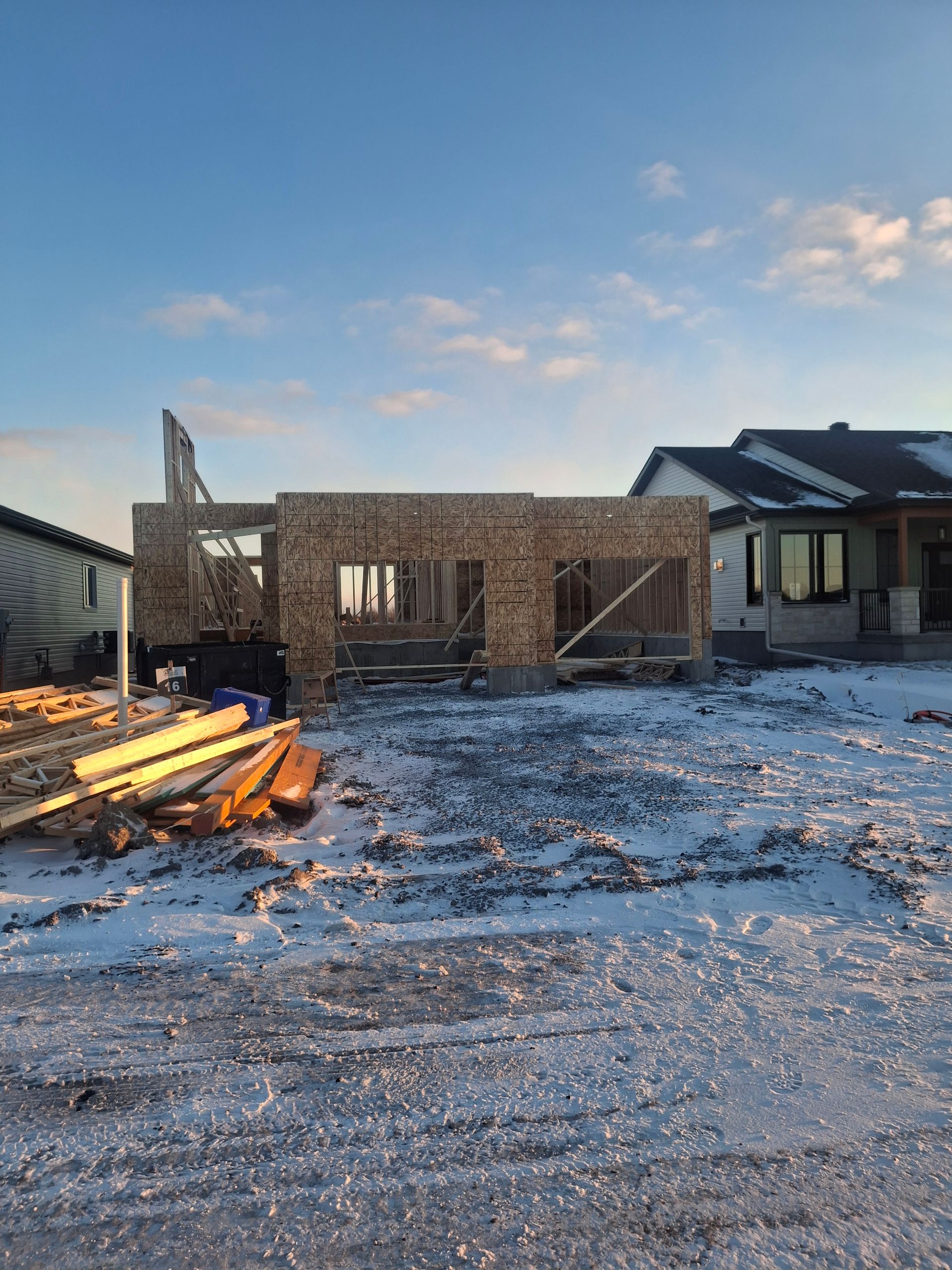Construction of a building's frame; wood and OSB on a snowy lot, another house in the background, blue sky.