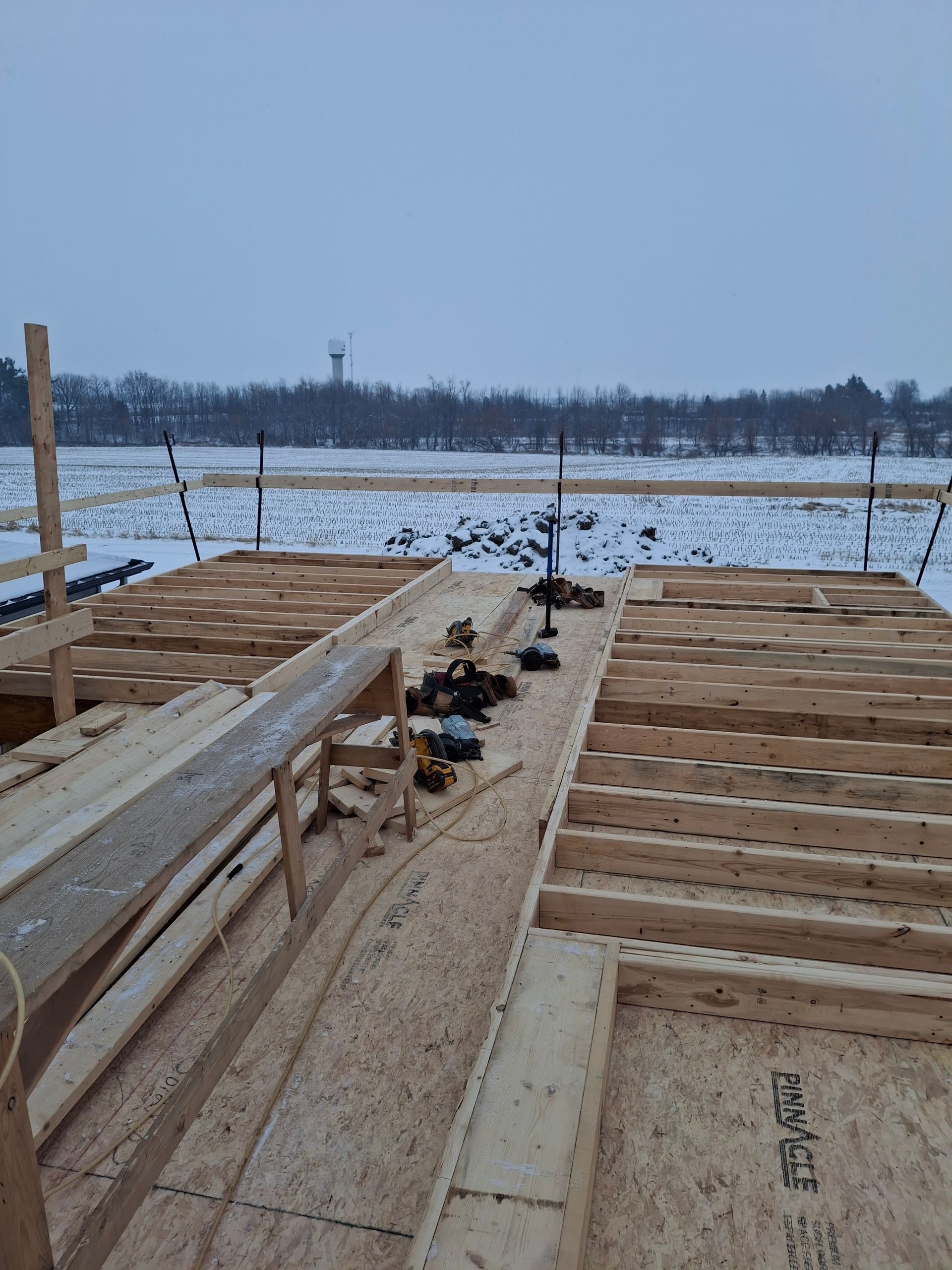 Construction site with wood framing on a snowy field, tools visible. Distant water tower.