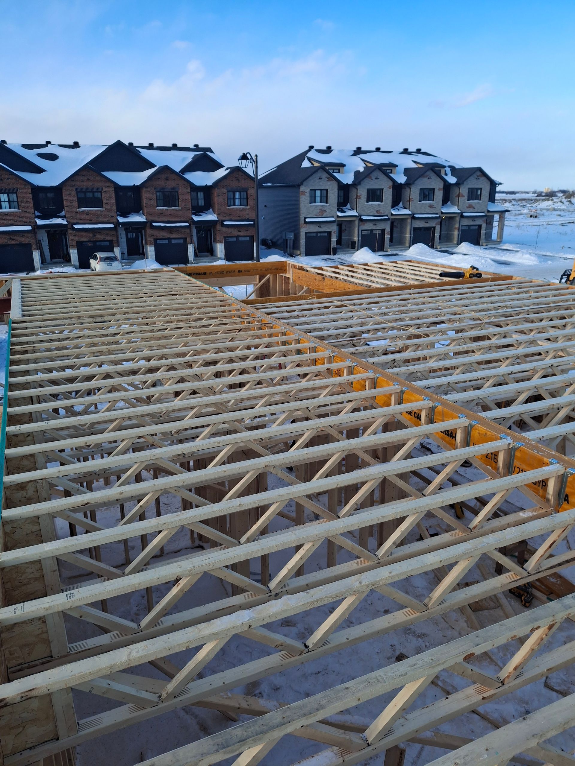 Wooden truss roof construction site with townhouses in the background on a snowy day.