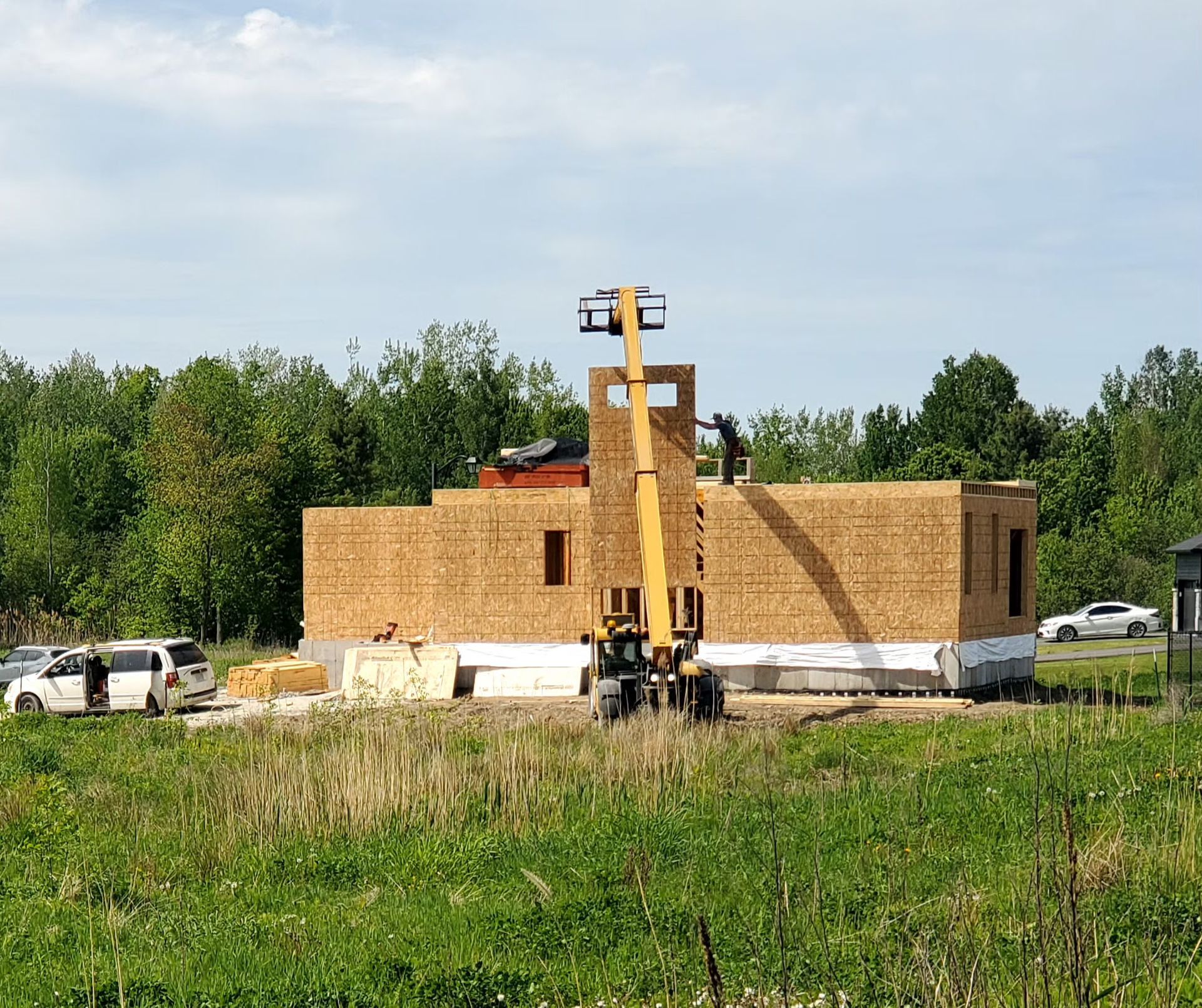 Construction site; a one-story building frame with a telehandler lifting materials. Cars and trees in the background.