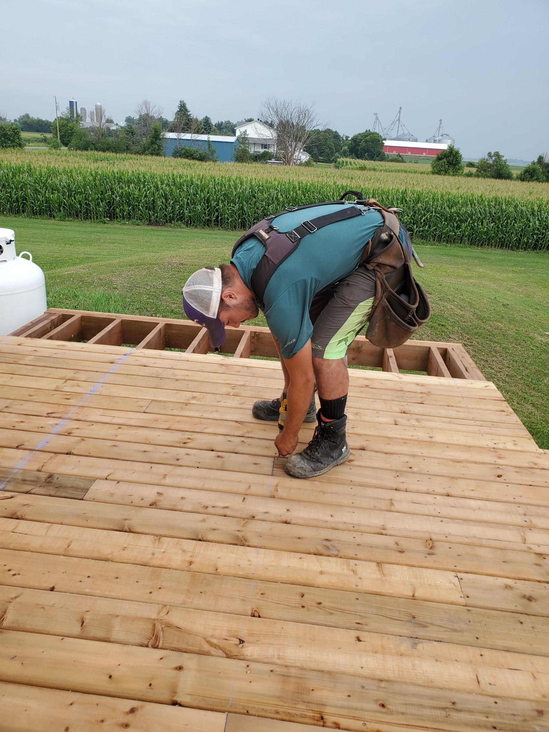 Carpenter installing wooden planks on a deck, outdoors. Wearing work gear and bending over to work, surrounded by a rural setting.