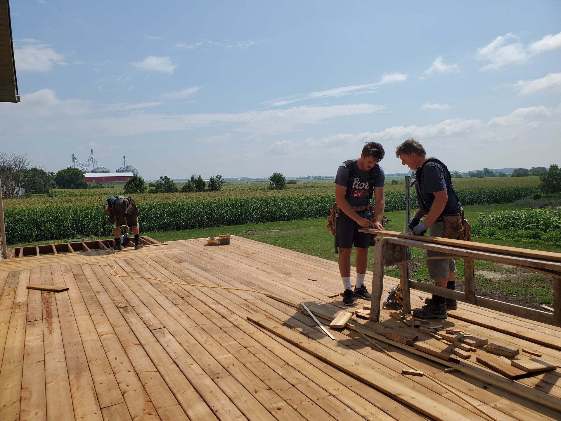 Three people building a wooden deck outdoors, surrounded by farmland. Sunny day.