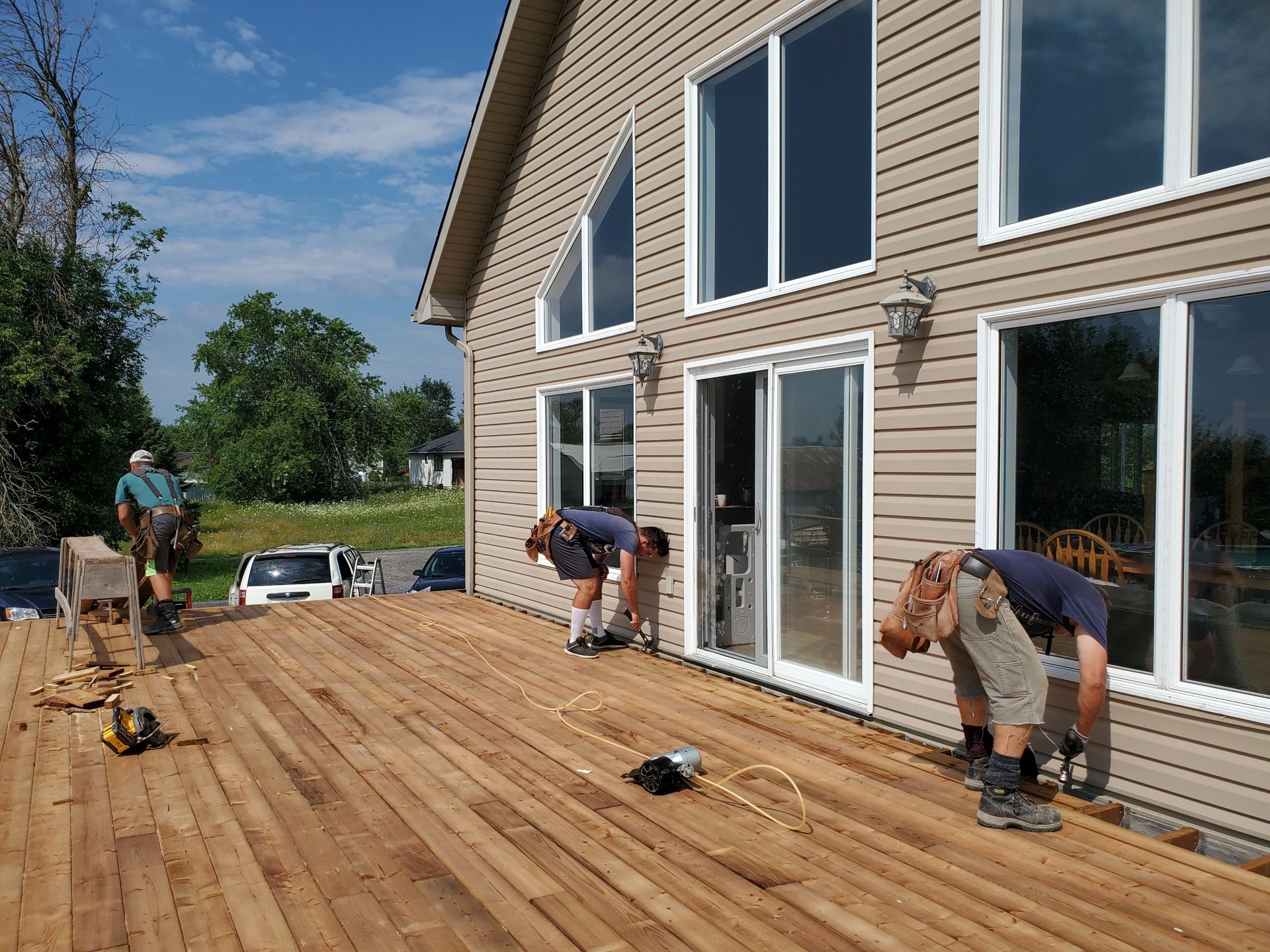 Three workers install decking on a wooden deck attached to a light brown sided house with large windows.