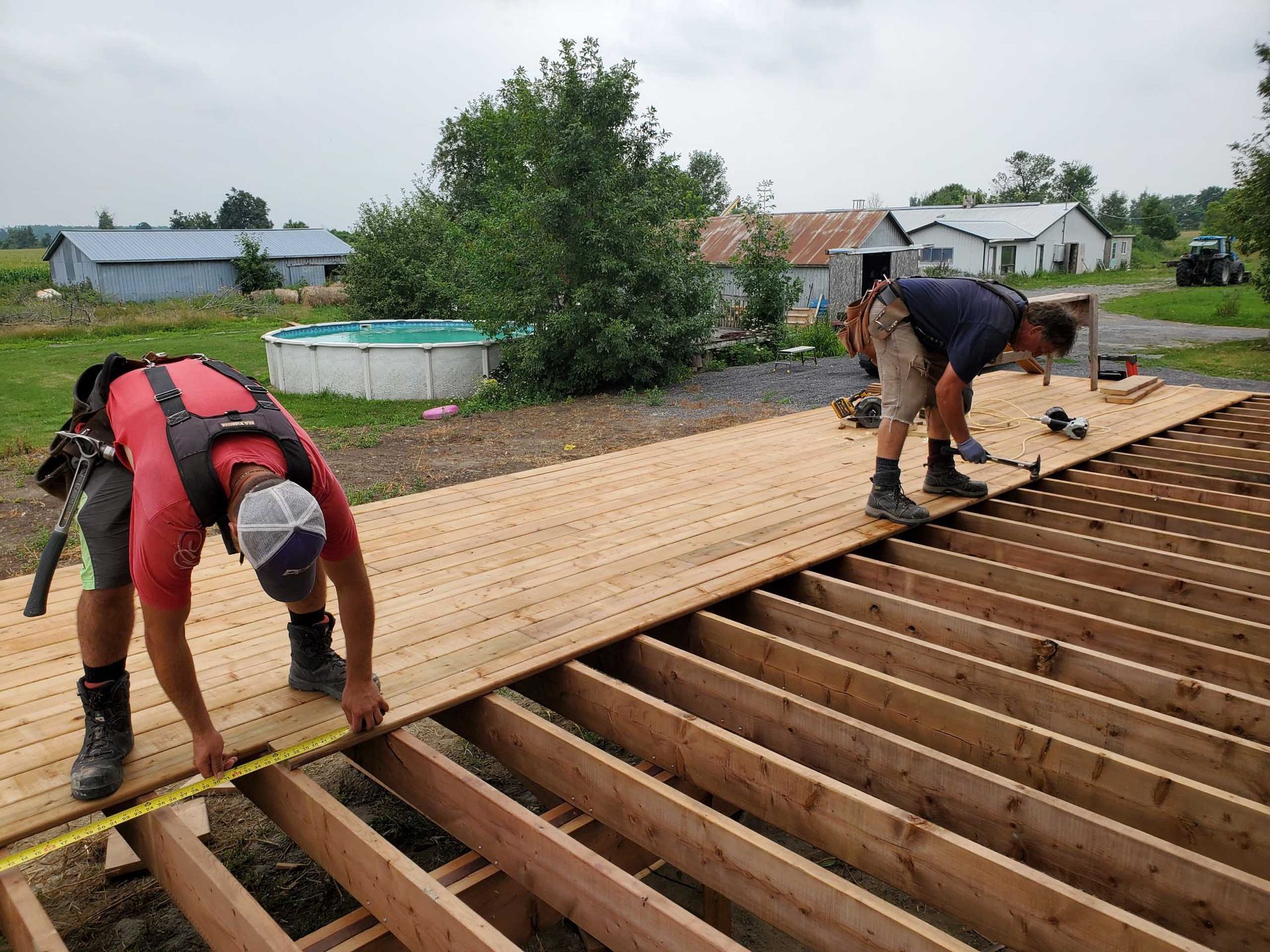 Two workers installing wood deck boards on a wood frame outdoors near a pool and buildings.