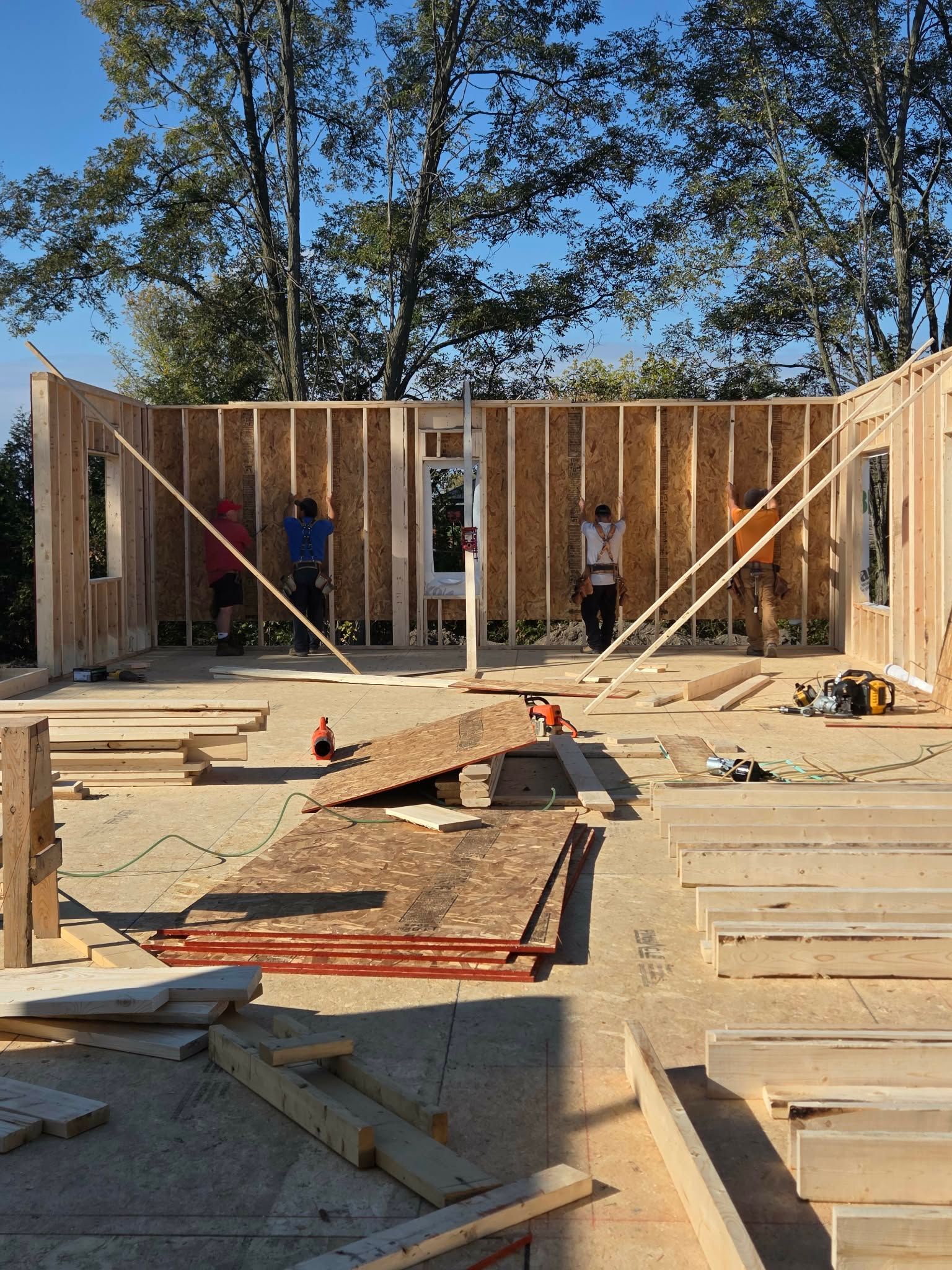 Construction workers framing a building's walls outdoors. Brown wood, blue sky, and tools on the ground.