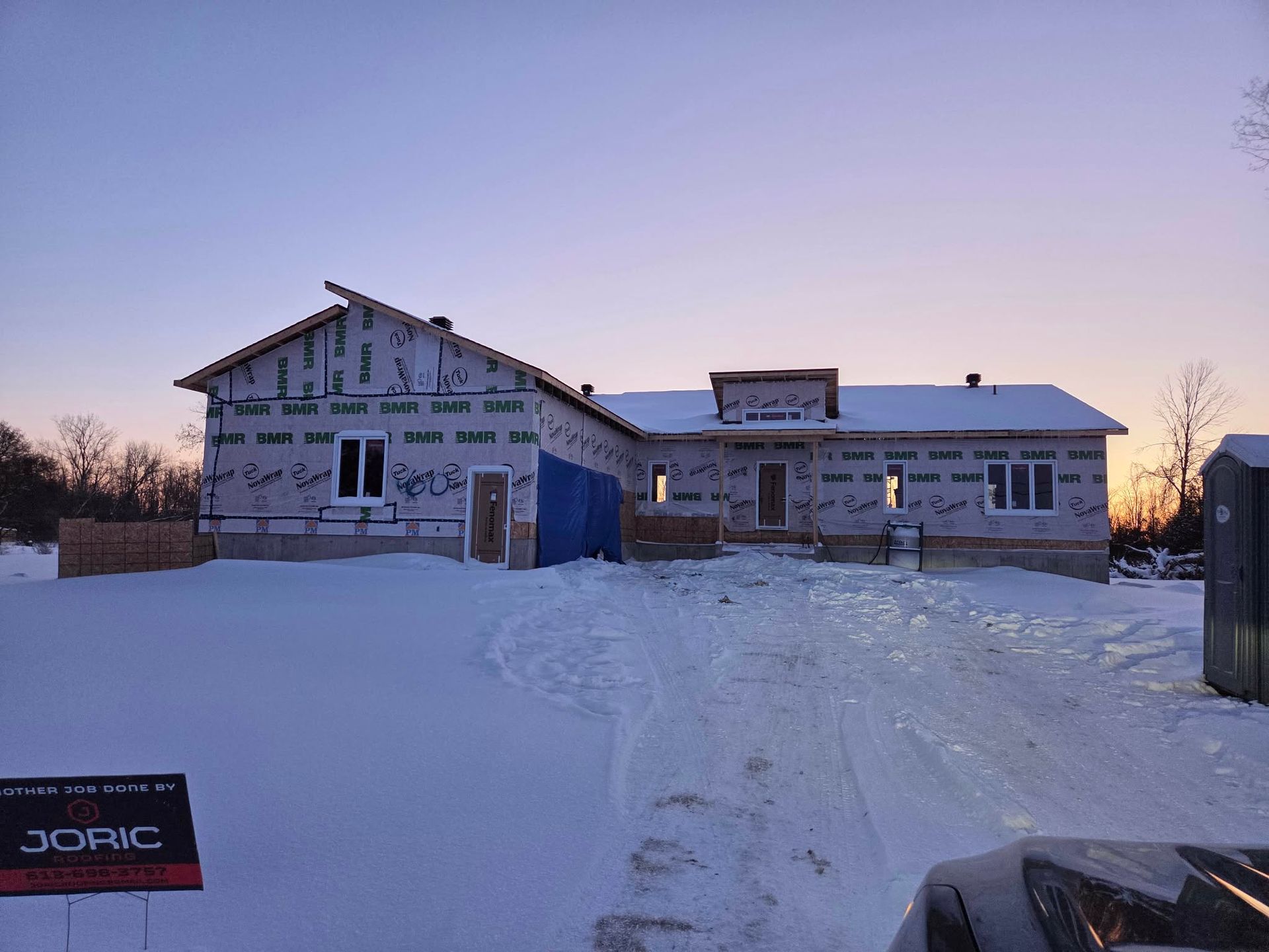 House under construction covered in weather barrier, set in a snow-covered landscape at dusk.