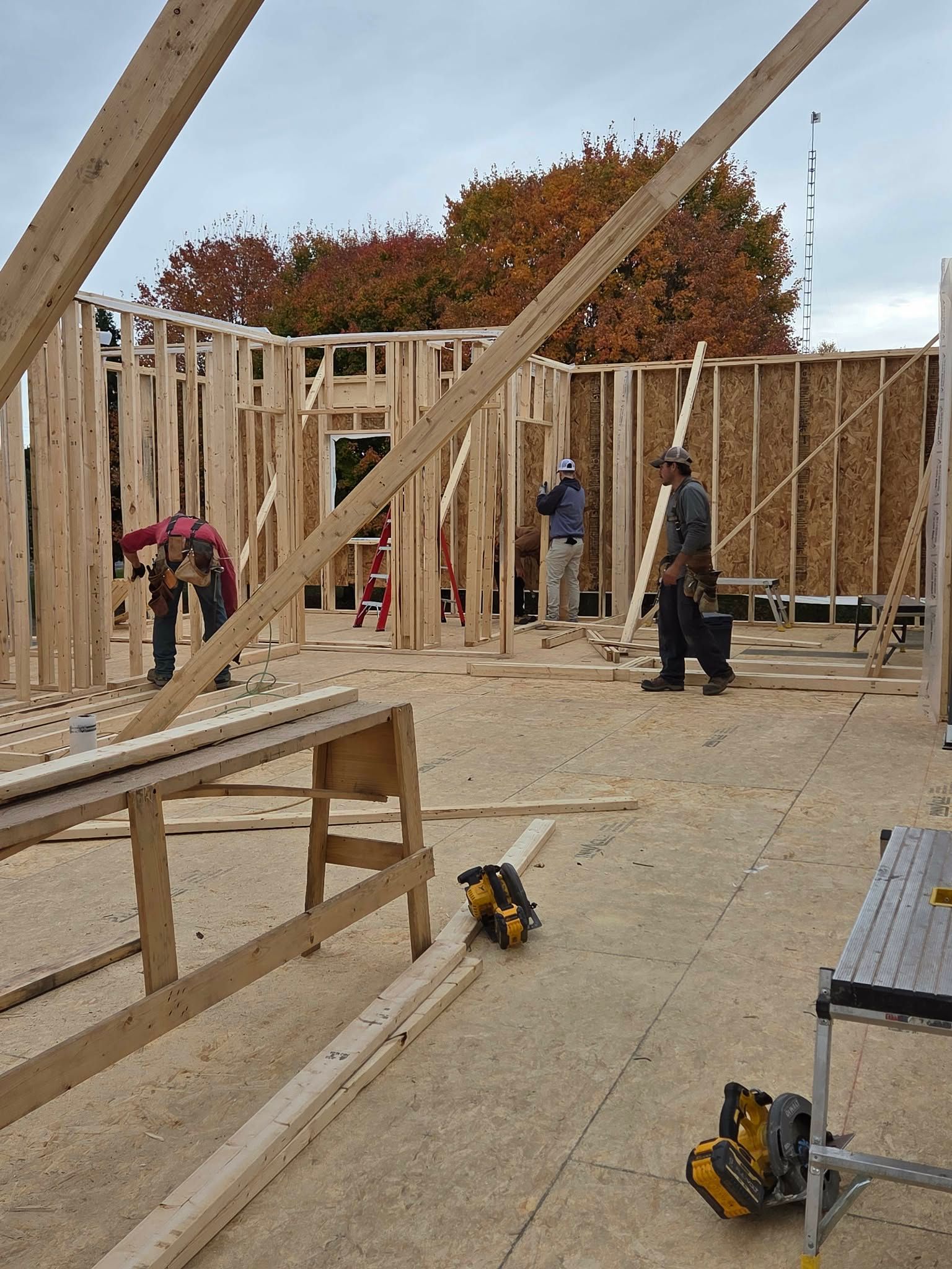 Construction workers framing a building. Wood beams, plywood, saws, and scaffolding on-site. Autumn trees in the background.