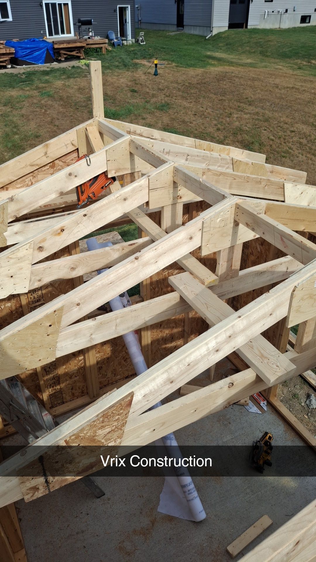 Wooden roof frame under construction, outdoors. Lumber pieces assembled, orange saw visible, sunny.