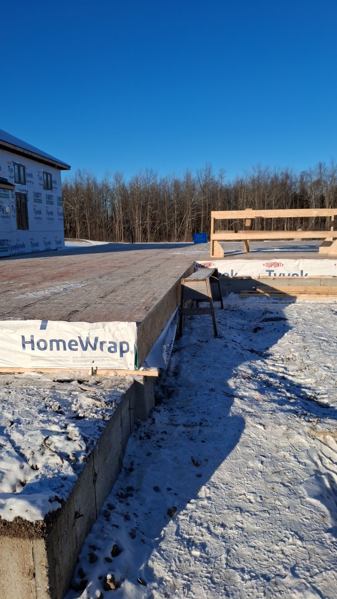 Construction site with a concrete foundation, covered with snow, and building materials; clear blue sky.